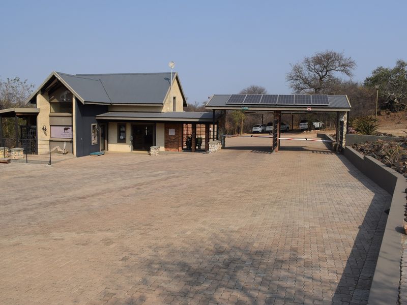 Front view of a residential estate entrance featuring a paved driveway, security gatehouse with solar panels, and stone masonry accents.