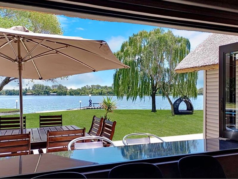 A view from a covered patio overlooking a lush green lawn and a serene lake, featuring a thatched roof structure and outdoor entertainment space.