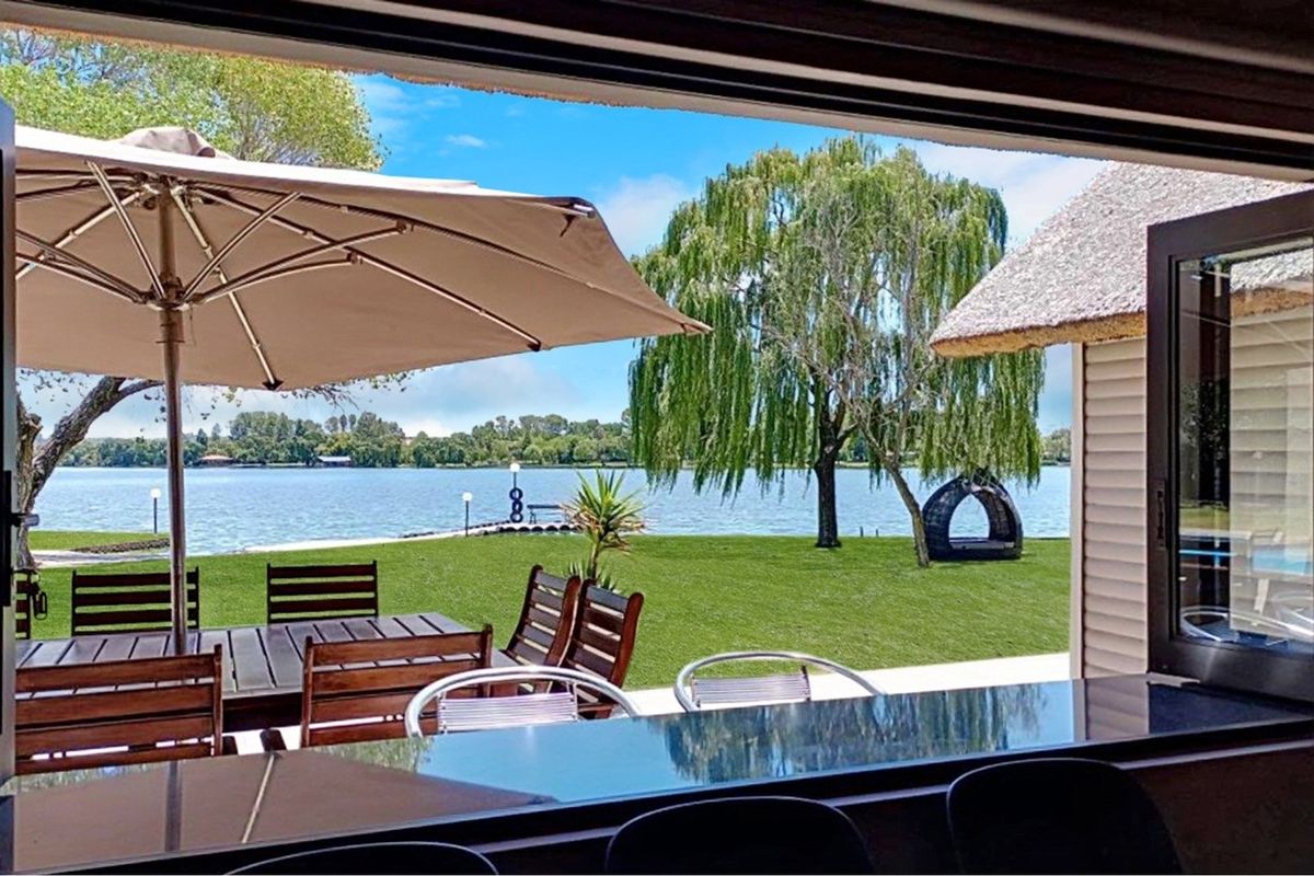 A view from a covered patio overlooking a lush green lawn and a serene lake, featuring a thatched roof structure and outdoor entertainment space.