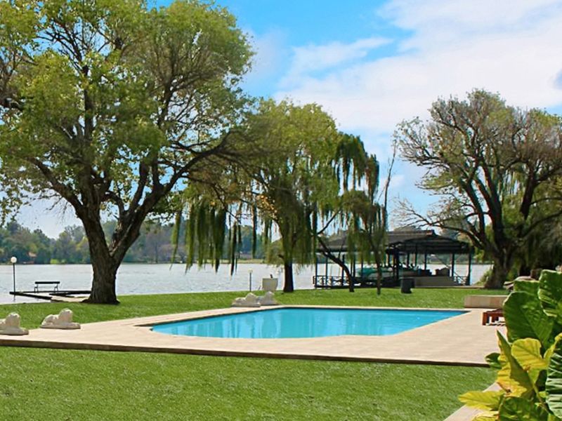 A scenic outdoor pool area overlooking a large dam, featuring a paved pool deck, expansive lawn, and a private jetty with a boat house.