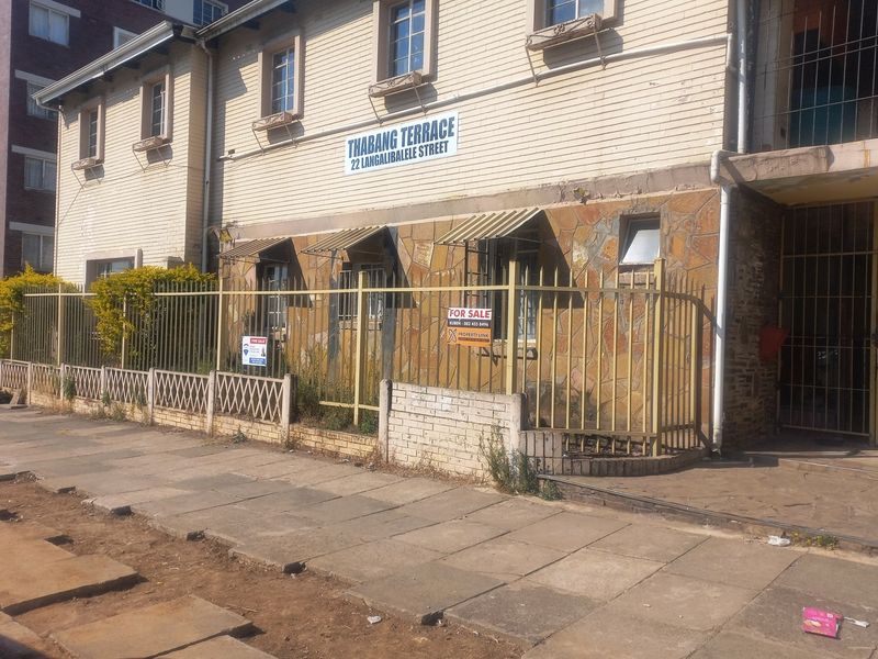 Front exterior view of Thabang Terrace apartment building featuring stone cladding, metal security fencing, and window awnings along a paved street.