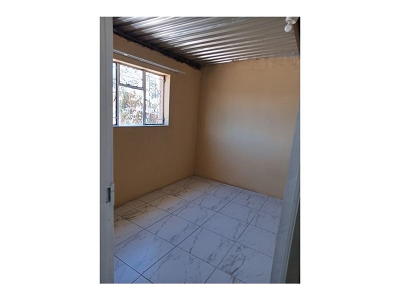 A small, empty bedroom featuring tiled flooring, a corrugated metal ceiling, and a window with security burglar bars.