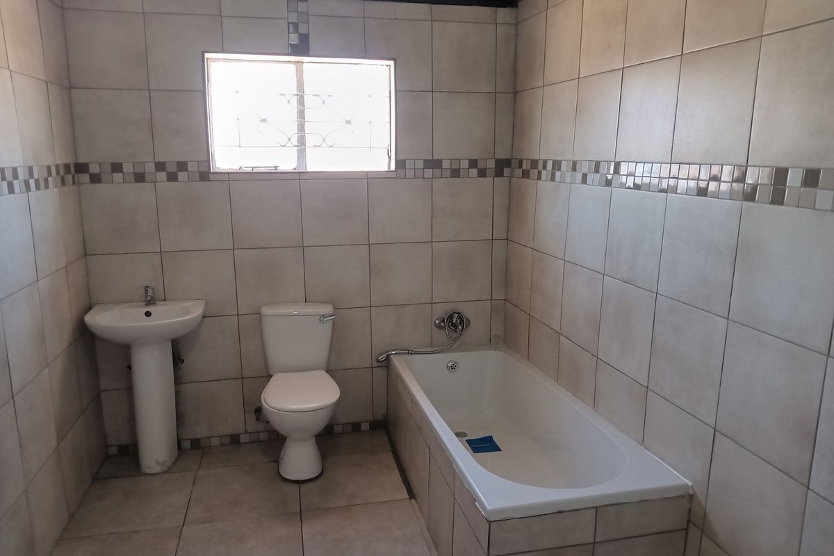 A functional bathroom featuring a pedestal basin, toilet, and bathtub with full-height wall tiling and a window with security bars.