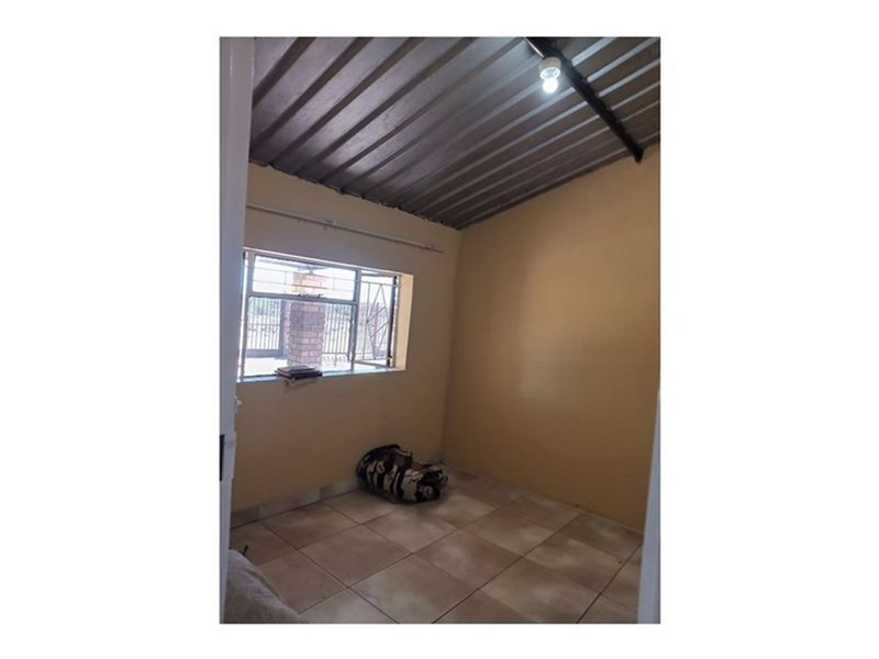 A simple, empty bedroom featuring tiled flooring, a corrugated metal ceiling, and a steel-framed window with security bars.
