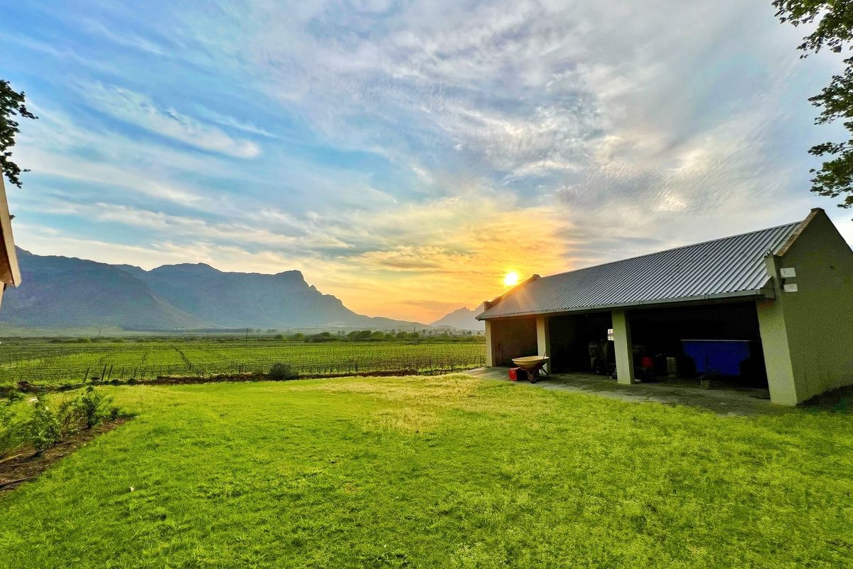 A scenic view of a farm property featuring a large open-sided barn structure, expansive green lawns, and a vineyard set against a mountain backdrop.