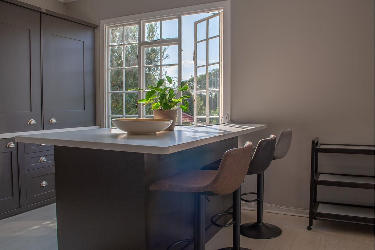 A modern kitchen featuring a dark-toned central island with integrated seating space, positioned beneath a large, light-filled casement window.