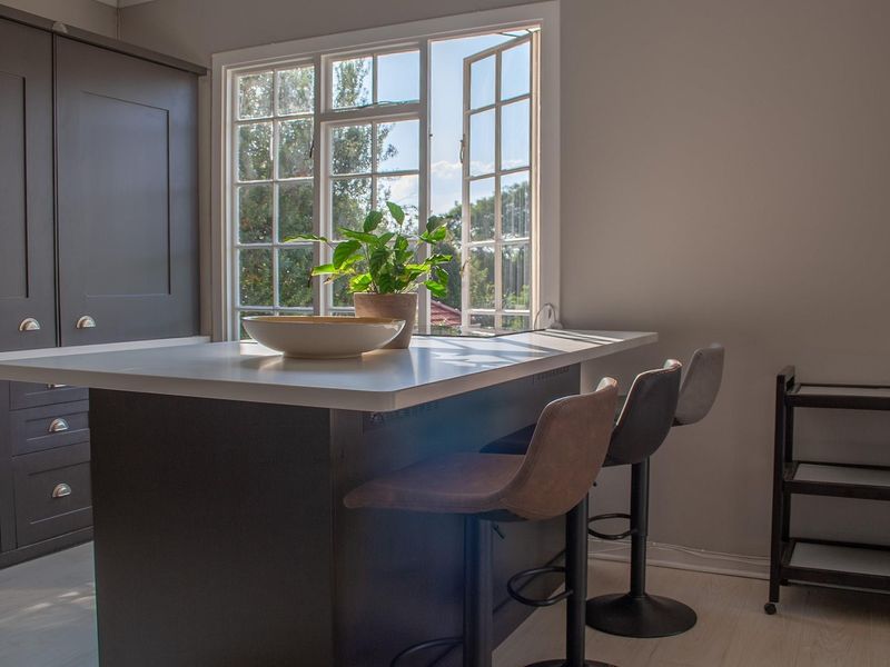 A modern kitchen featuring a dark-toned central island with integrated seating space, positioned beneath a large, light-filled casement window.