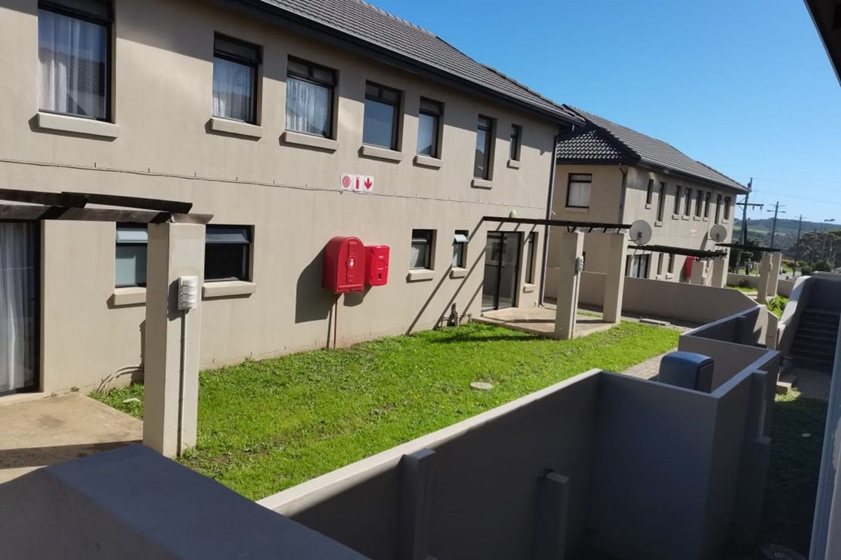 Rear exterior view of the apartment complex featuring a shared lawn area, paved patios with pergolas, and installed fire safety equipment.