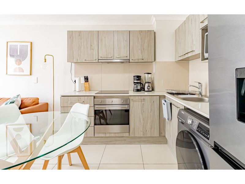 Modern open-plan kitchen featuring light wood-grain cabinetry, integrated stainless steel oven, electric hob, extractor fan, and tiled flooring.