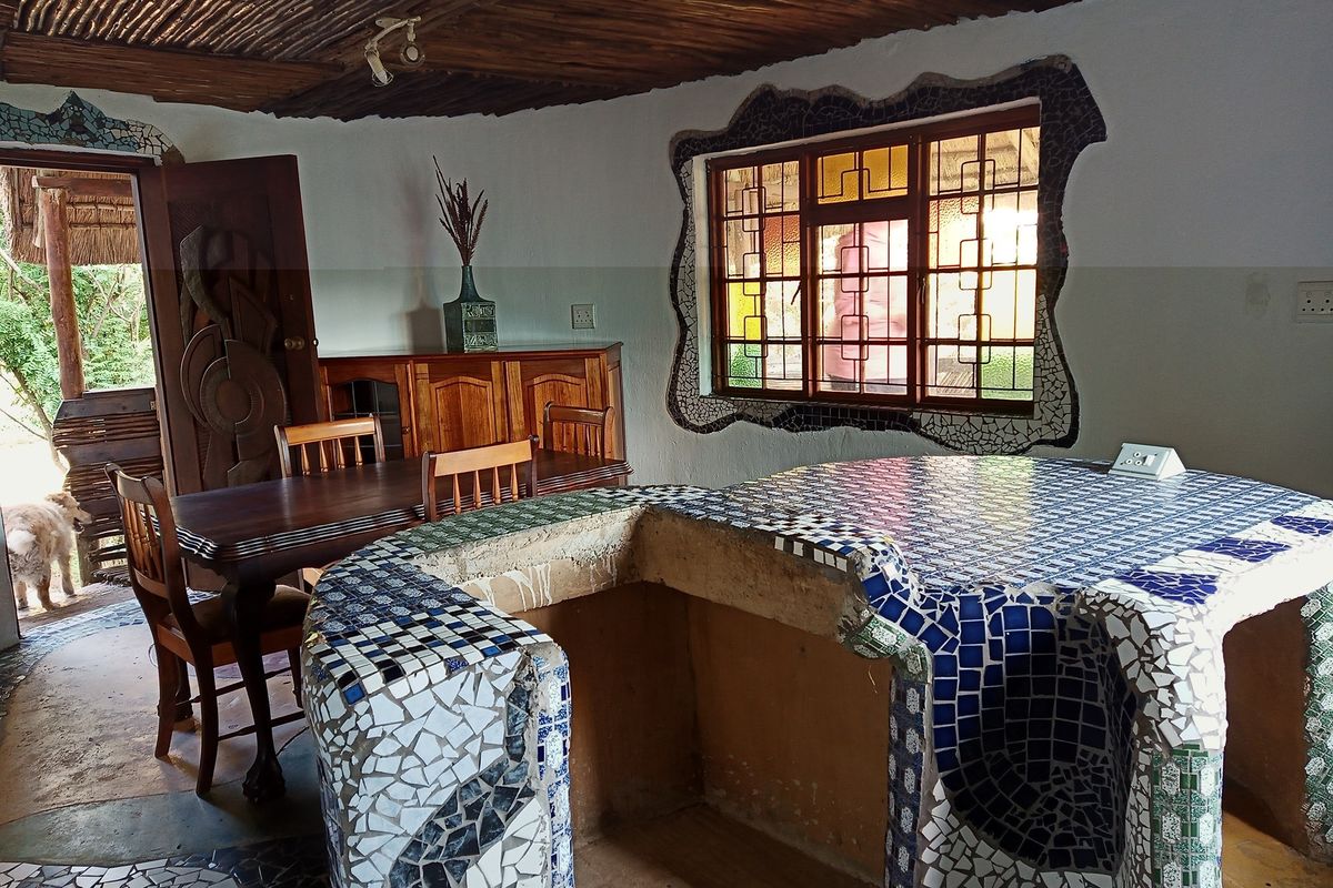 A rustic kitchen area featuring a unique mosaic-tiled island, a traditional thatched ceiling, and a distinctive mosaic-framed window.