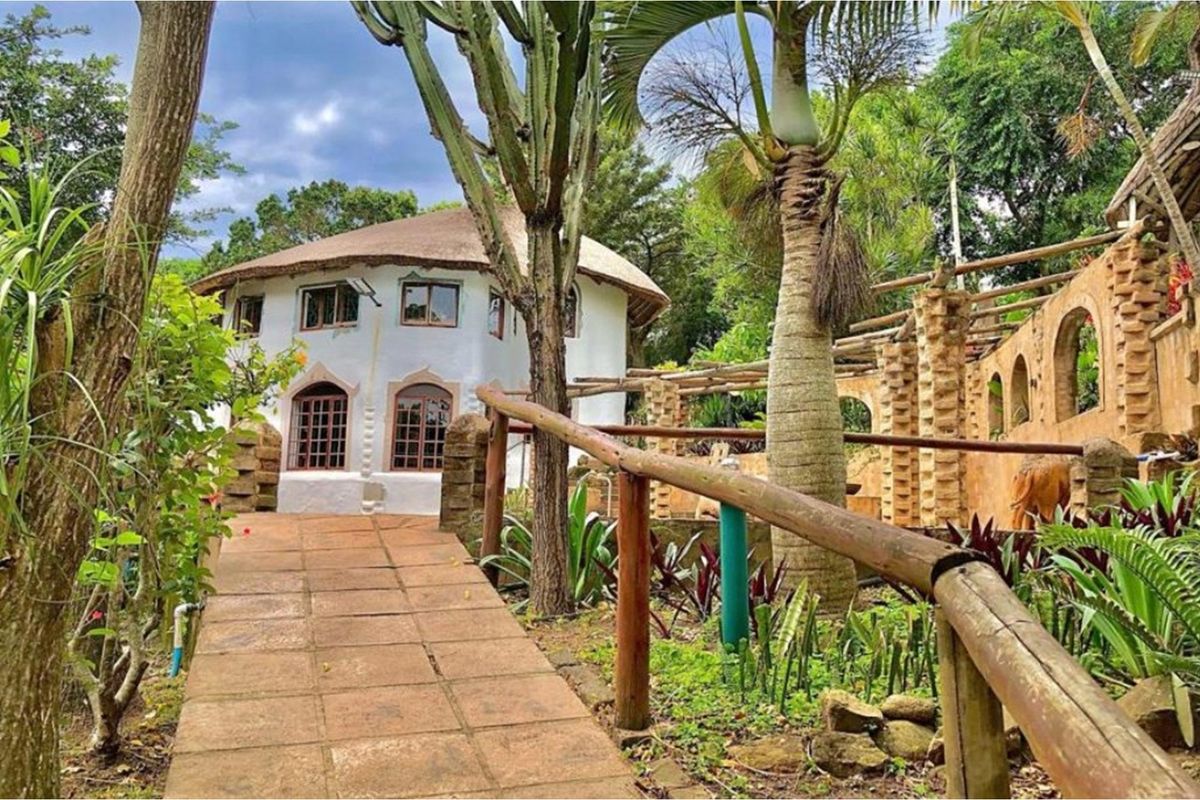 A unique circular dwelling with a thatched roof, featuring a stone-paved walkway and rustic brick architectural elements set within a lush garden environment.