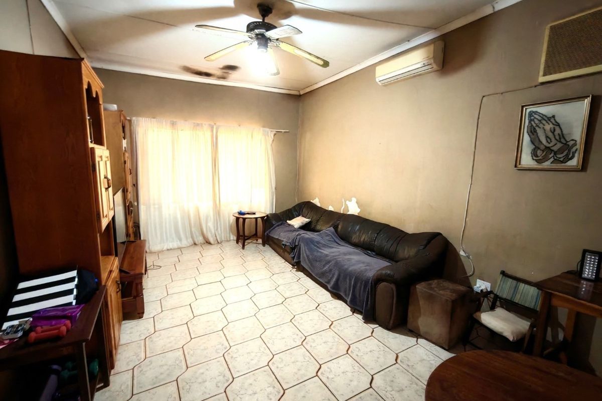 A living room featuring tiled flooring, a ceiling fan, and wall-mounted air conditioning unit, showing signs of wear and maintenance requirements.