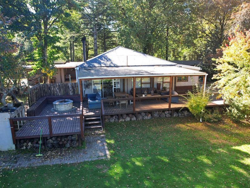 Elevated view of a rear property featuring a spacious timber deck, built-in fire pit area, and a covered patio overlooking a lush garden setting.