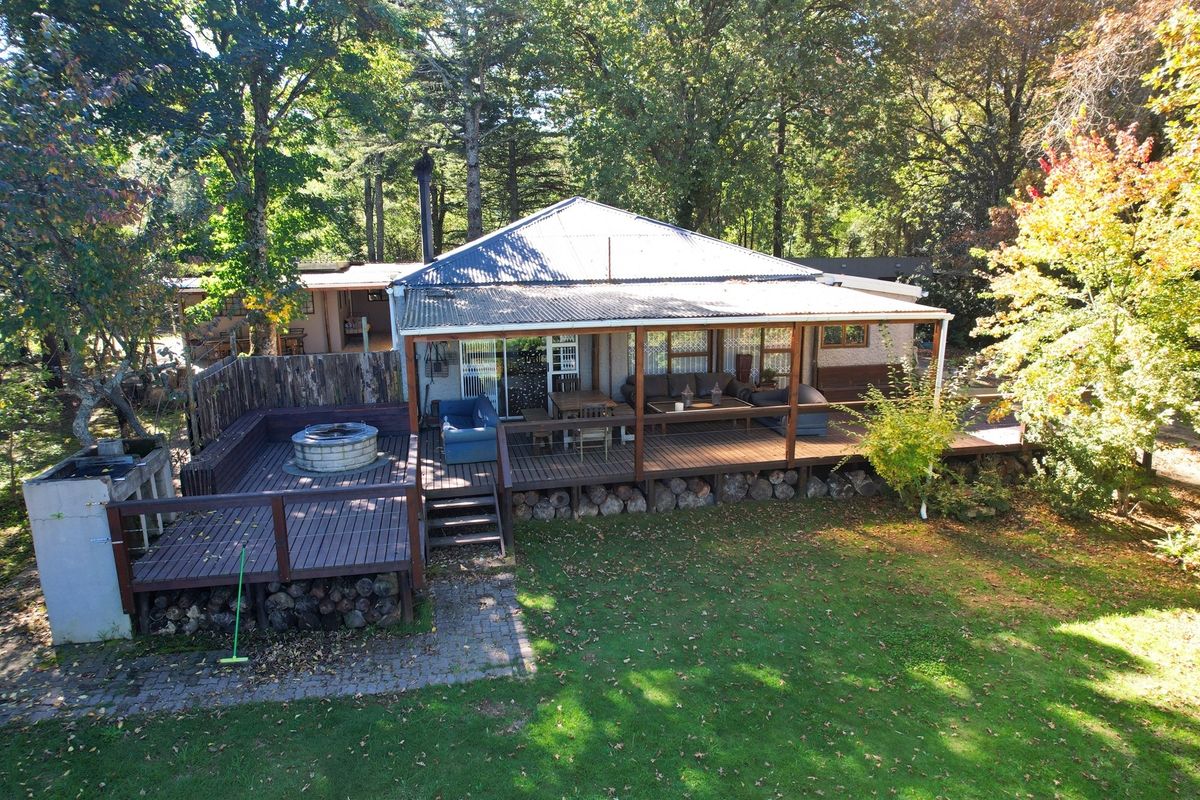 Elevated view of a rear property featuring a spacious timber deck, built-in fire pit area, and a covered patio overlooking a lush garden setting.