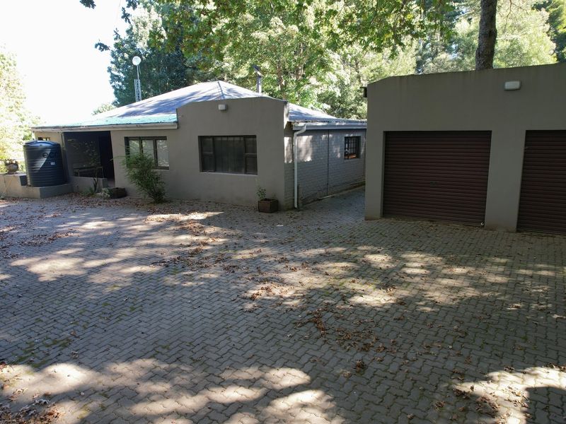 Exterior view of the property featuring a paved driveway, double garage, and main house structure surrounded by mature trees.