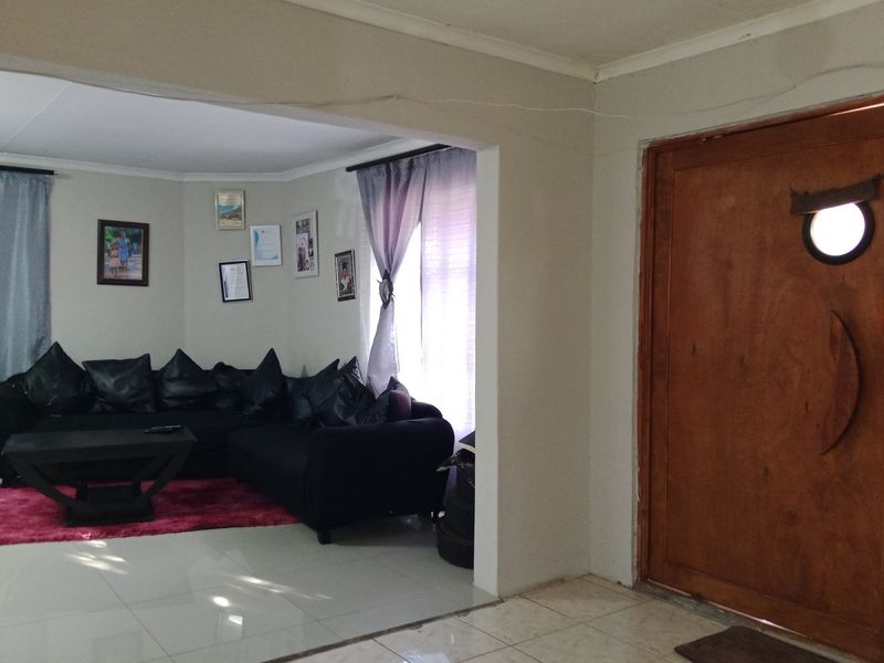 A living room area featuring tiled flooring and a wooden entrance door, with an open-plan layout connecting to the main lounge space.