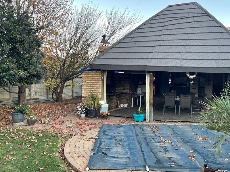 Outdoor entertainment area featuring a covered patio with built-in brick braai, adjacent to a swimming pool with a safety cover and garden space.