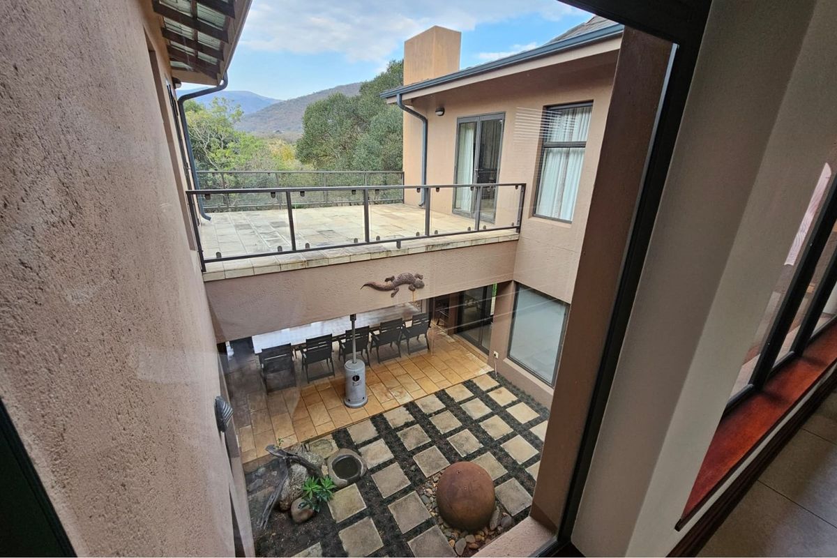An elevated view of a rear courtyard featuring a paved patio area, a balcony with glass railings, and landscaped garden elements.