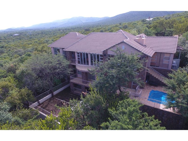 Aerial view of a multi-level residential property featuring stone cladding, tiled roofing, multiple balconies, and a private swimming pool set in a natural landscape.