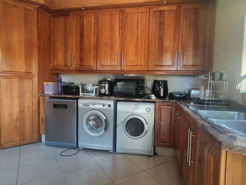 Functional scullery featuring extensive wooden cabinetry, granite countertops, a double stainless steel sink, and tiled flooring with dedicated space for laundry appliances.