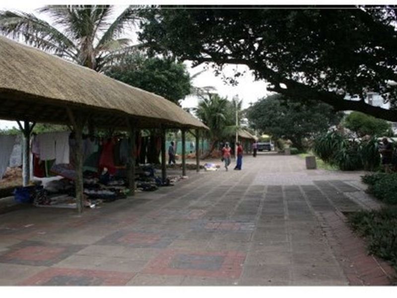 An outdoor communal area featuring a long thatched-roof structure and paved walkway, surrounded by mature trees and landscaping.