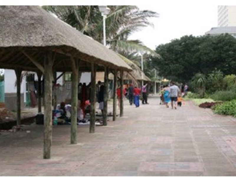 A paved outdoor walkway featuring a series of rustic thatched-roof shelters supported by wooden posts, situated in a landscaped garden area.
