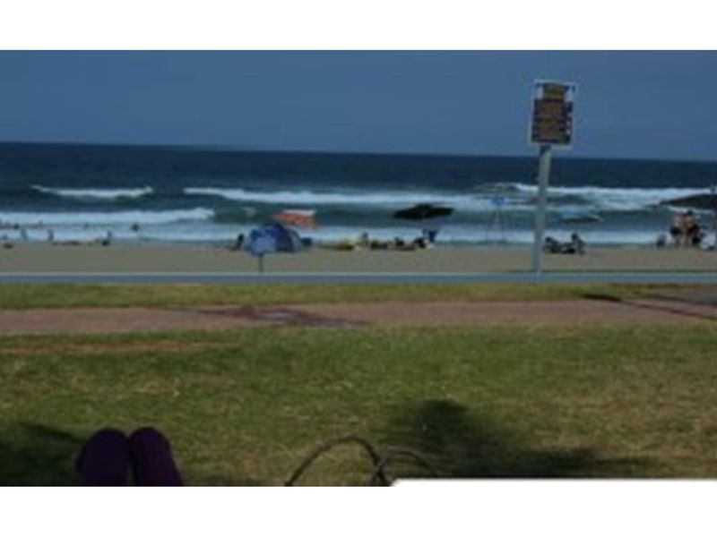 A view of the beach and ocean from a grassy public area, featuring a paved walkway and signage.