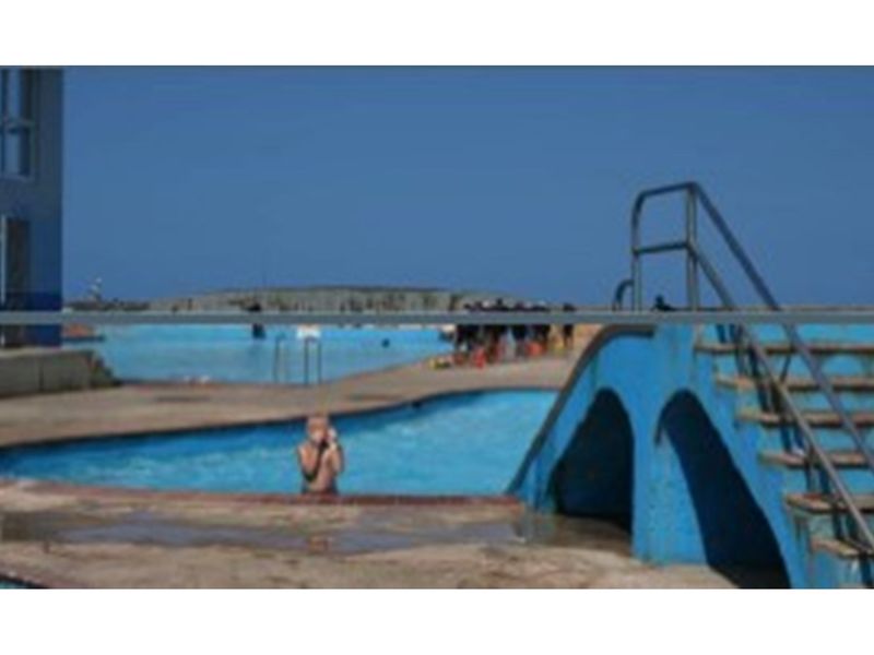 Outdoor swimming pool area featuring a concrete deck, blue painted stairs, and metal handrails, set against a coastal backdrop.