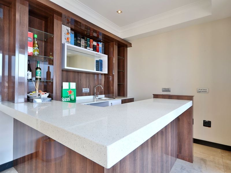 A modern built-in home bar featuring dark wood cabinetry, a stone countertop, integrated sink, and glass shelving with recessed lighting.