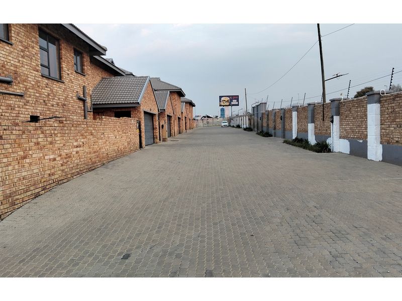 A view of the paved driveway and access road within a townhouse complex, featuring brick boundary walls with electric fencing and multiple garage entrances.