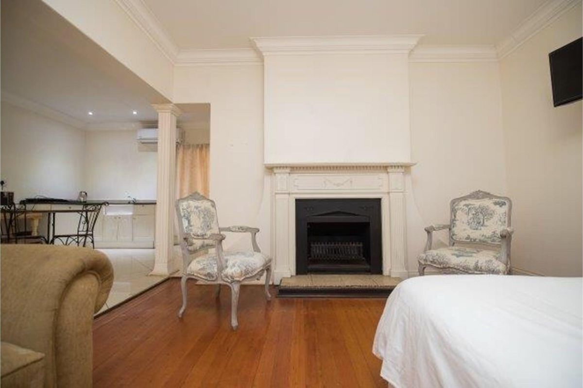 A living room featuring a classic fireplace with a mantelpiece, hardwood flooring, and crown molding, with an adjacent view into a kitchen area.