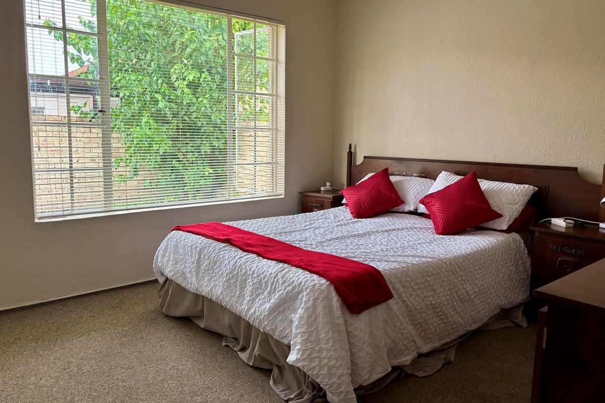 A standard bedroom featuring neutral wall tones, carpeted flooring, and a large window with fitted blinds providing natural light.