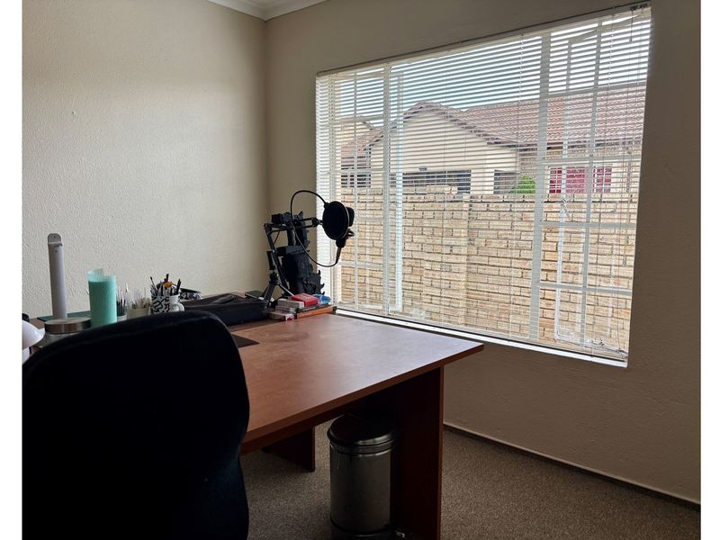 A functional home office space featuring neutral wall finishes, carpeted flooring, and a large window with fitted blinds overlooking the exterior boundary wall.