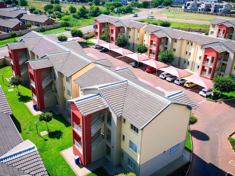 Aerial view of a modern apartment complex featuring multi-story residential buildings, paved parking areas with shade sails, and well-maintained communal garden spaces.
