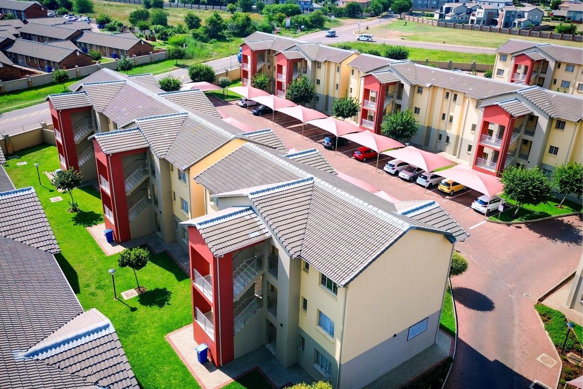 Aerial view of a modern apartment complex featuring multi-story residential buildings, paved parking areas with shade sails, and well-maintained communal garden spaces.