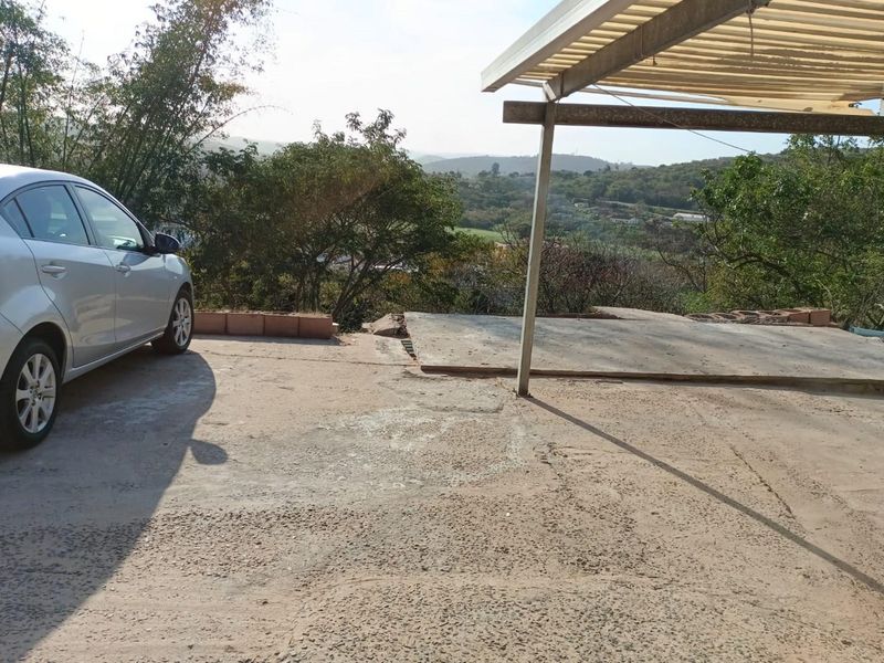 An outdoor driveway area featuring a basic carport structure and scenic hillside views, showing signs of wear on the concrete surface.
