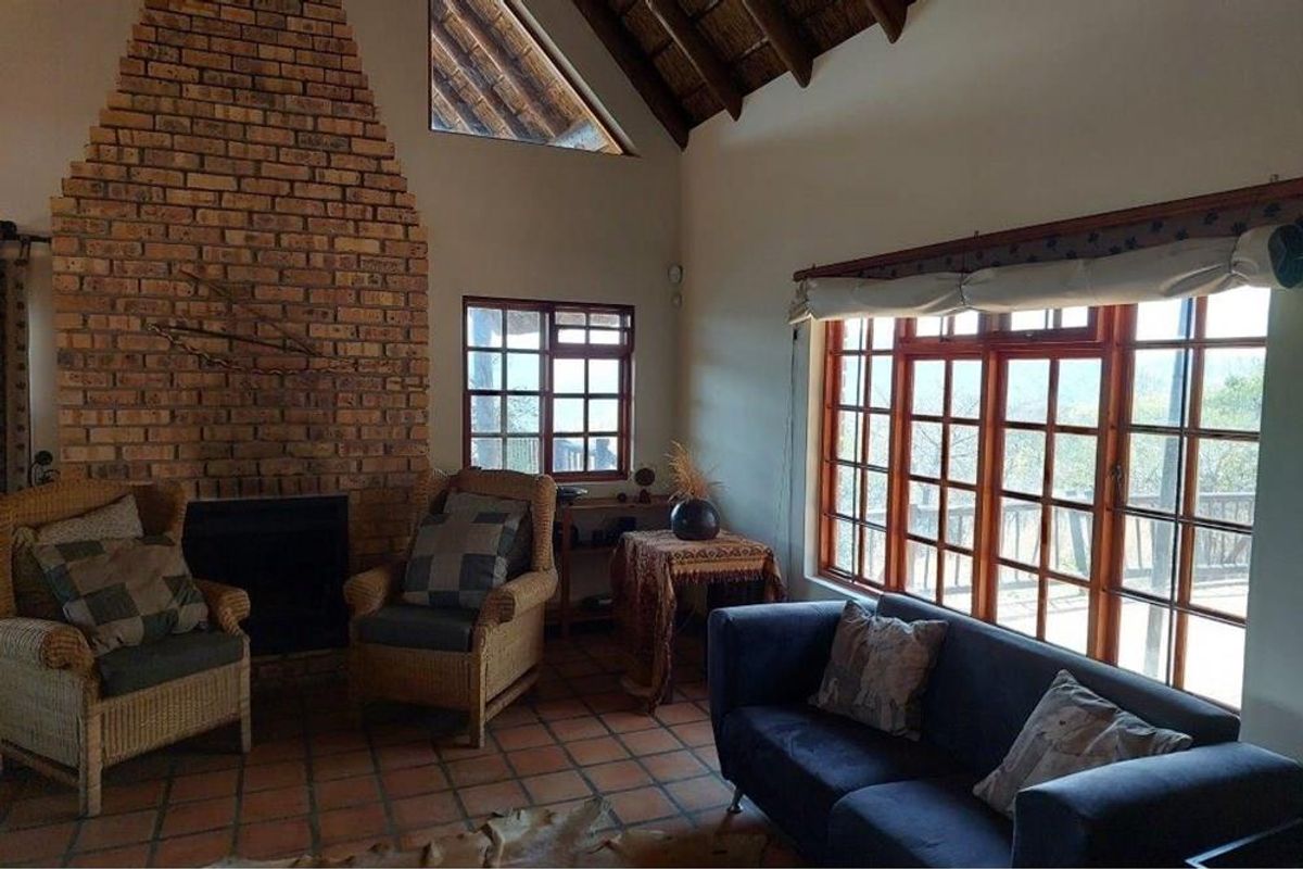 A cozy living room featuring a prominent exposed brick fireplace, vaulted thatched ceiling, and large timber-framed windows offering natural light.