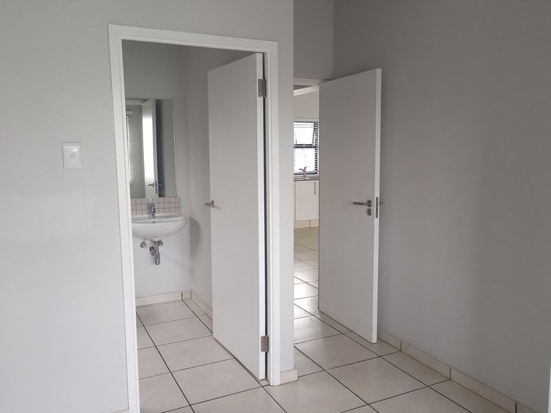 A view of a hallway leading to a bathroom and an adjacent room, featuring neutral wall tones and tiled flooring throughout.