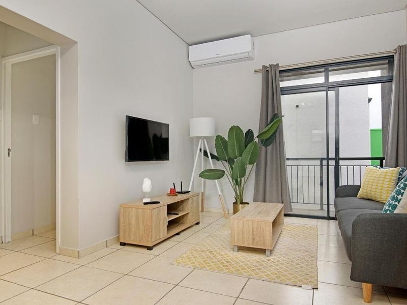 A well-maintained living room featuring tiled flooring, a wall-mounted air conditioning unit, and direct access to a private balcony through sliding glass doors.