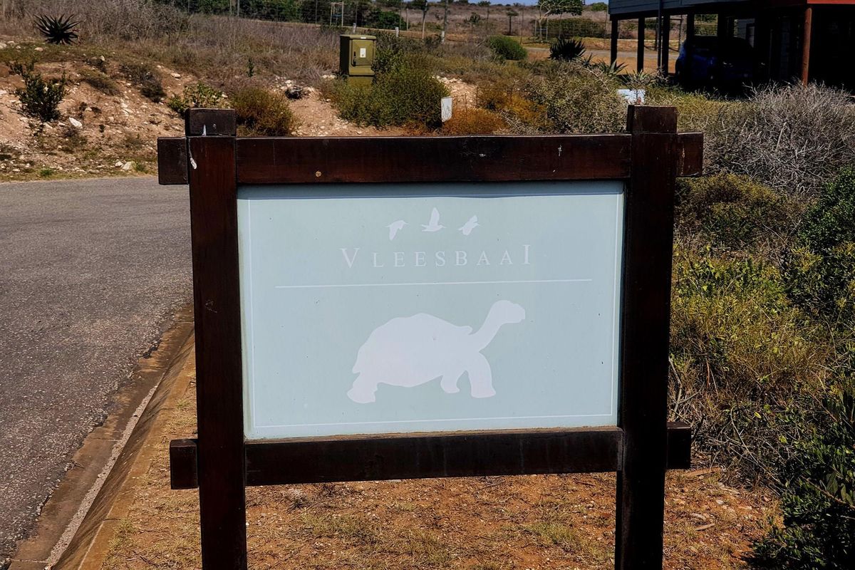 A signage board marking the Vleesbaai area, situated along a paved road with surrounding natural coastal vegetation.