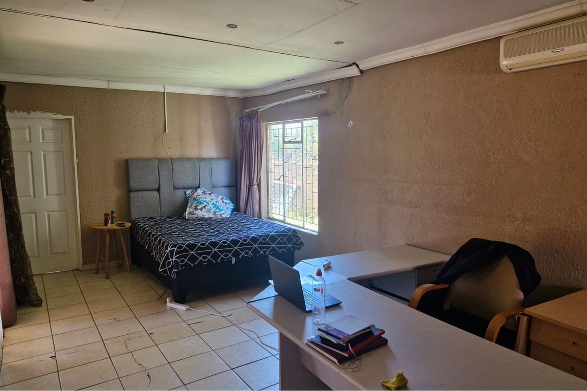 A bedroom featuring tiled flooring, a wall-mounted air conditioning unit, and security bars on the window, requiring cosmetic updates to address wall and floor defects.