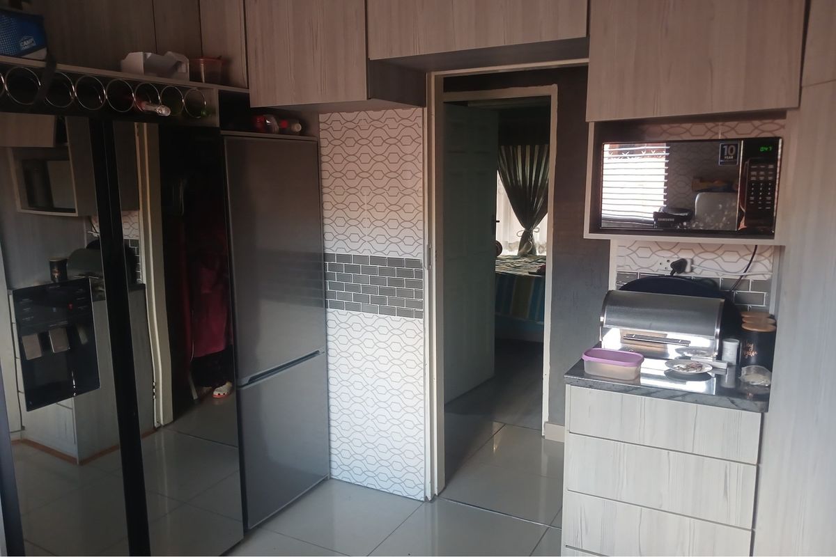 A functional kitchen area featuring light wood-grain cabinetry, tiled splashbacks, and a built-in microwave, with a doorway leading to an adjacent bedroom.