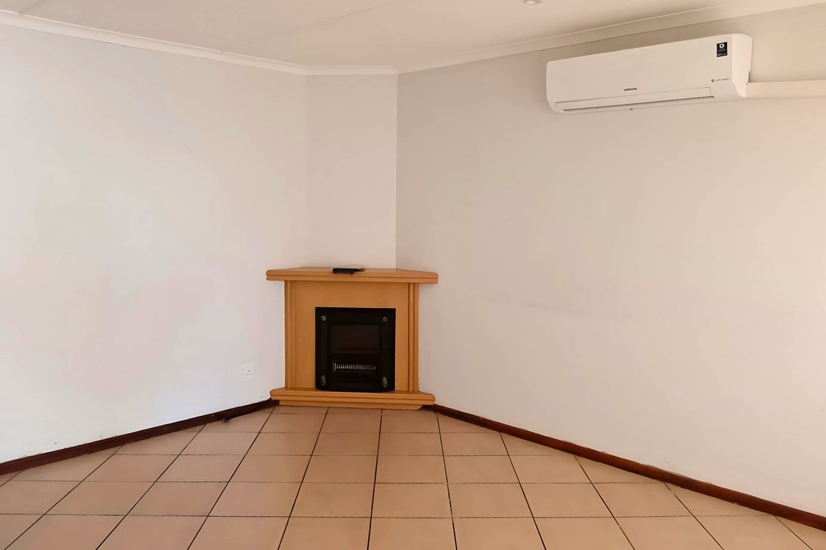 A bright, empty living room featuring tiled flooring, a corner fireplace, and a wall-mounted air conditioning unit.