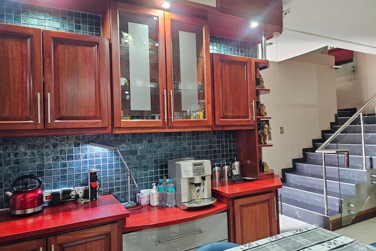 A kitchen area featuring rich wooden cabinetry, red countertops, and a dark mosaic tile backsplash, adjacent to a staircase with a metal railing.