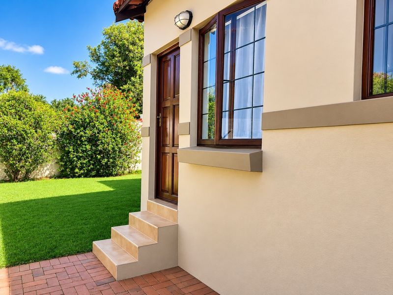 A well-maintained rear exterior view featuring a timber door, tiled steps, brick paving, and a neat garden area.