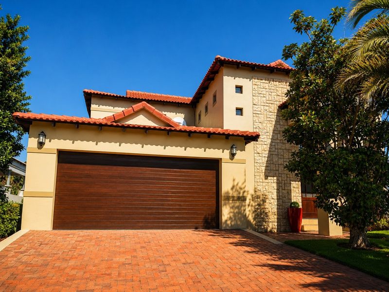 A well-maintained front elevation featuring a double garage, stone-clad accents, terracotta roof tiles, and a neat brick-paved driveway.