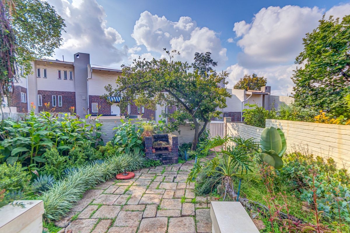 A private rear garden featuring a brick-paved patio area with a built-in brick braai, surrounded by established garden beds and boundary walls.