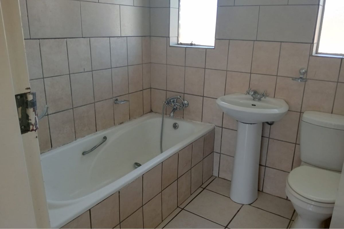A dated bathroom featuring a built-in bathtub with shower mixer, pedestal basin, and toilet, finished with neutral wall and floor tiling.