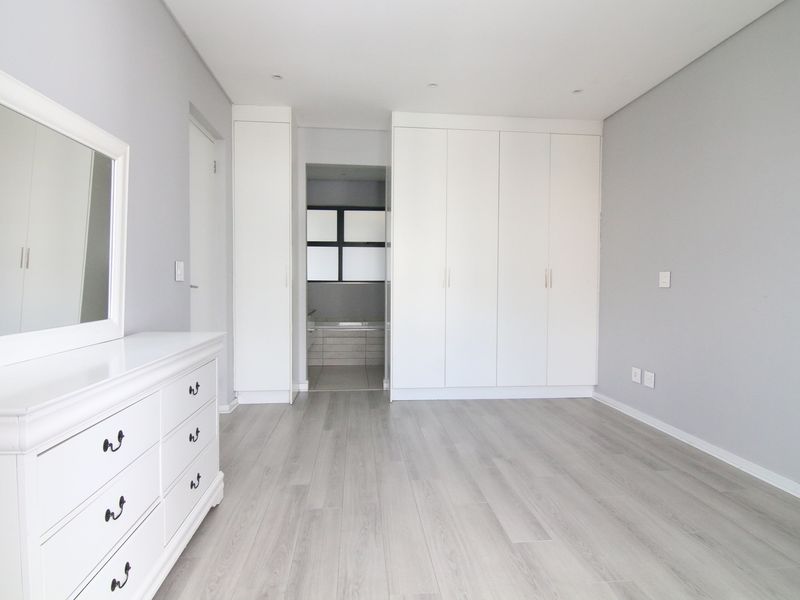 A bright, modern bedroom featuring light wood-look flooring, built-in white cabinetry, and an en-suite bathroom entrance.