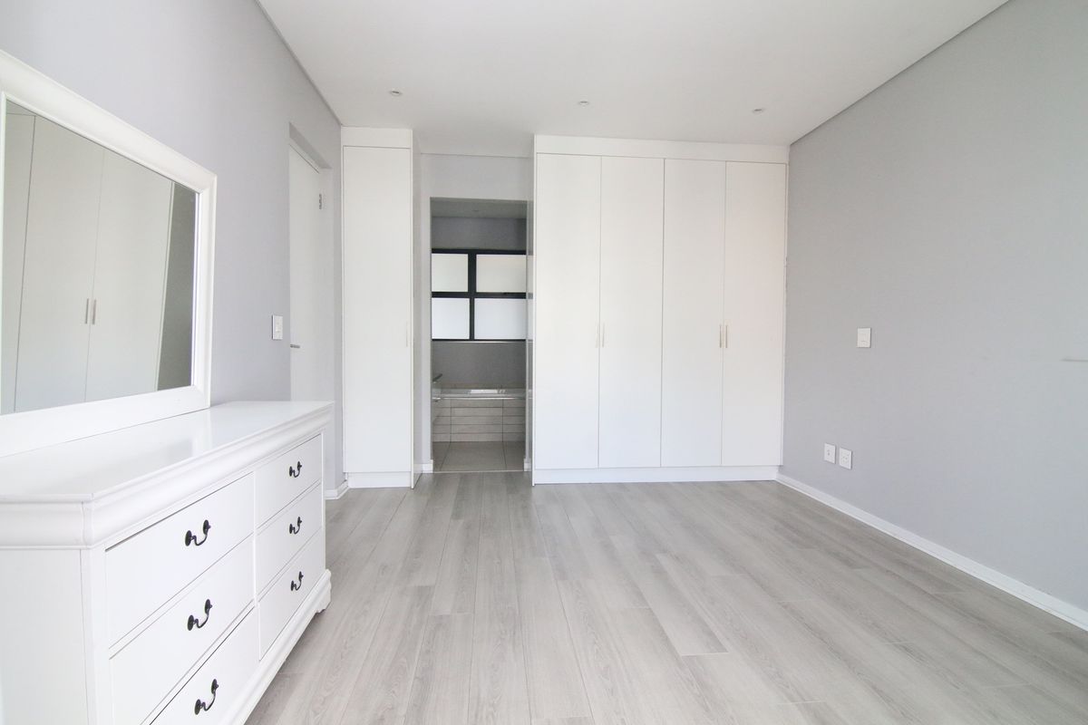 A bright, modern bedroom featuring light wood-look flooring, built-in white cabinetry, and an en-suite bathroom entrance.
